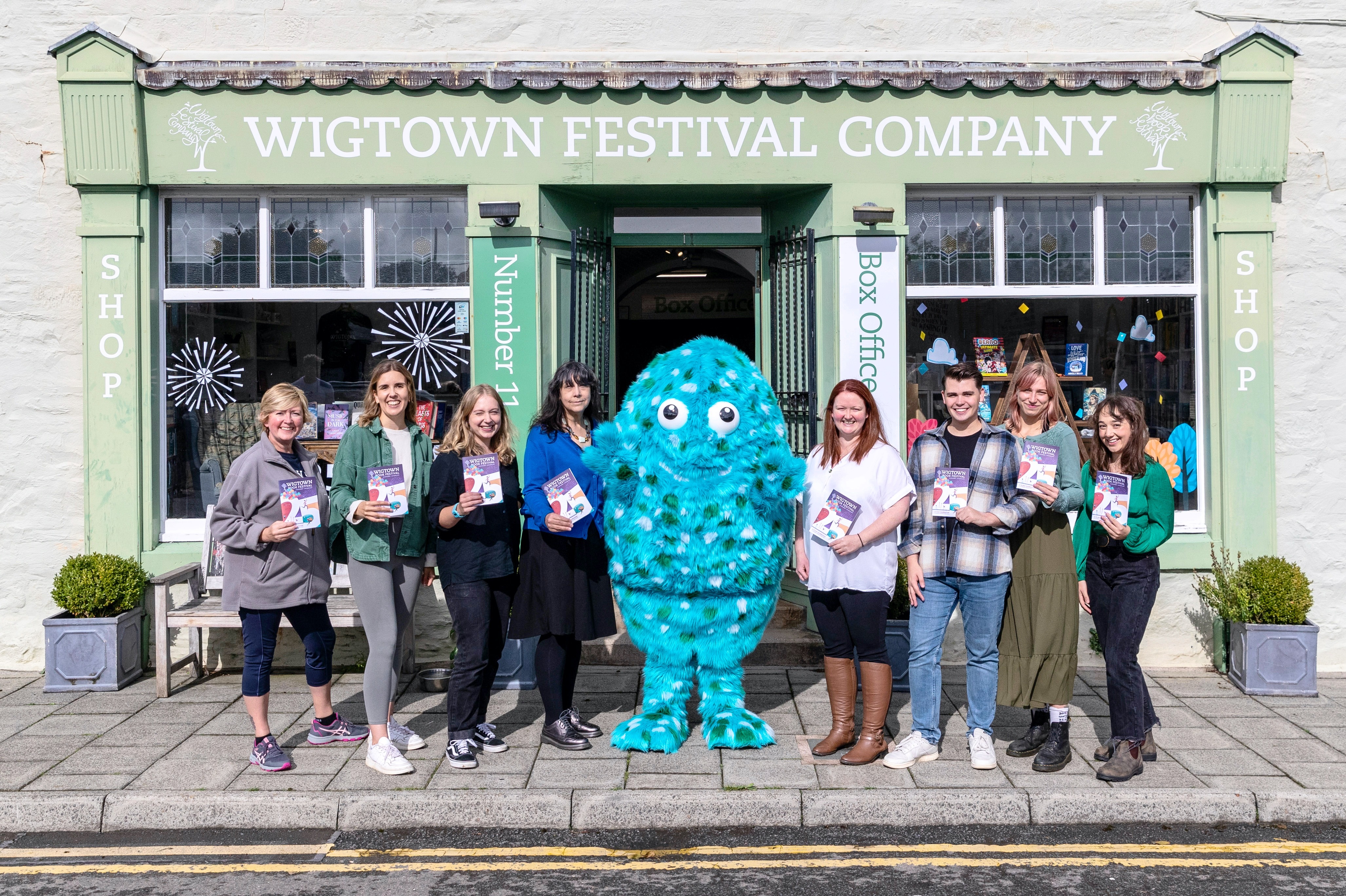 Eight staff members stand either side of Big Wig, the big blue book festival mascot, outside the number 11 bookshop. Each staff member is holding a 2023 festival brochure in front of them.