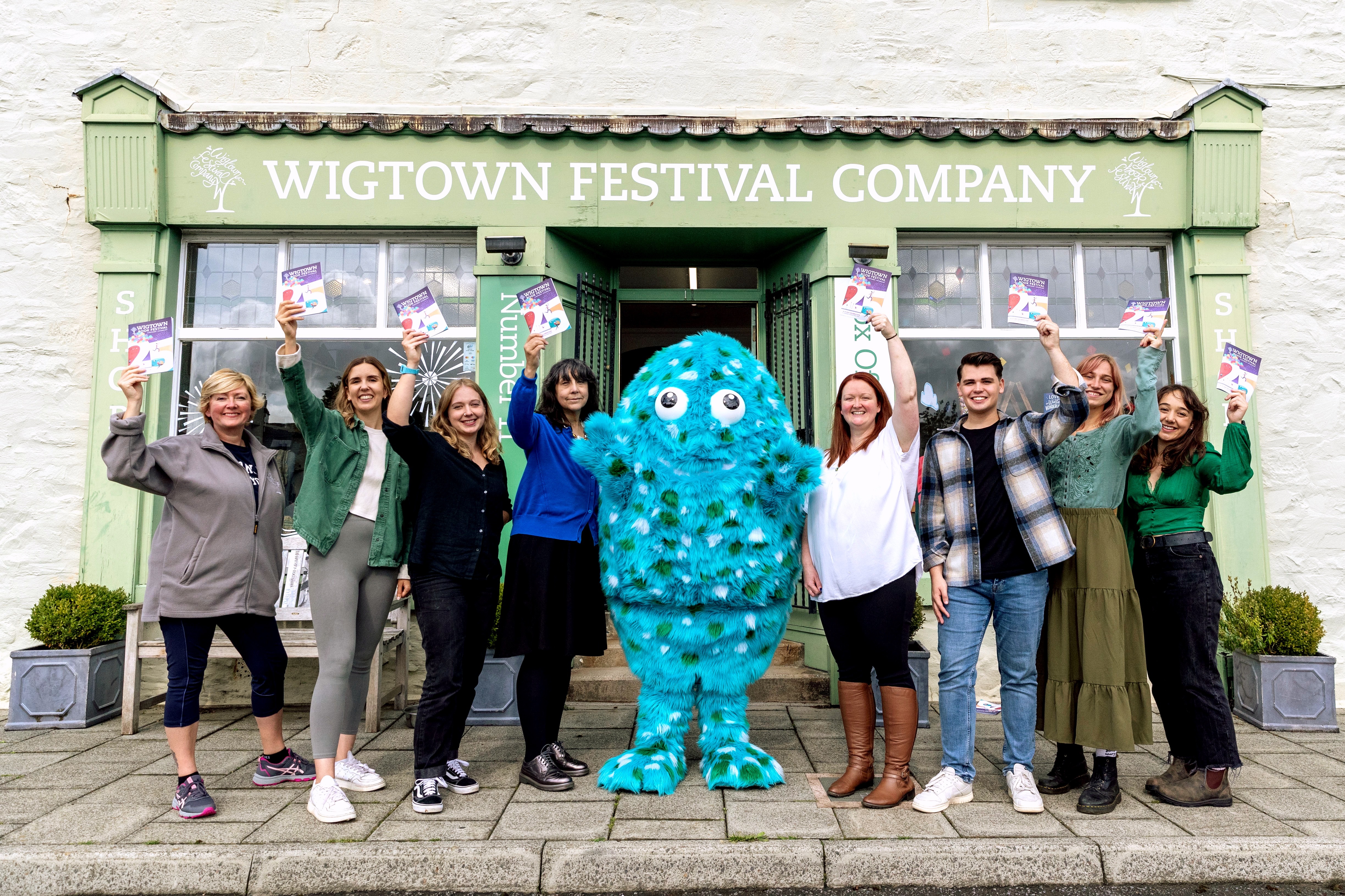 Eight staff members stand beside Big Wig, the big, blue, furry festival mascot, outside the Wigtown Book Festival bookshop. Each each staff holds up a 2023 programme brochure.