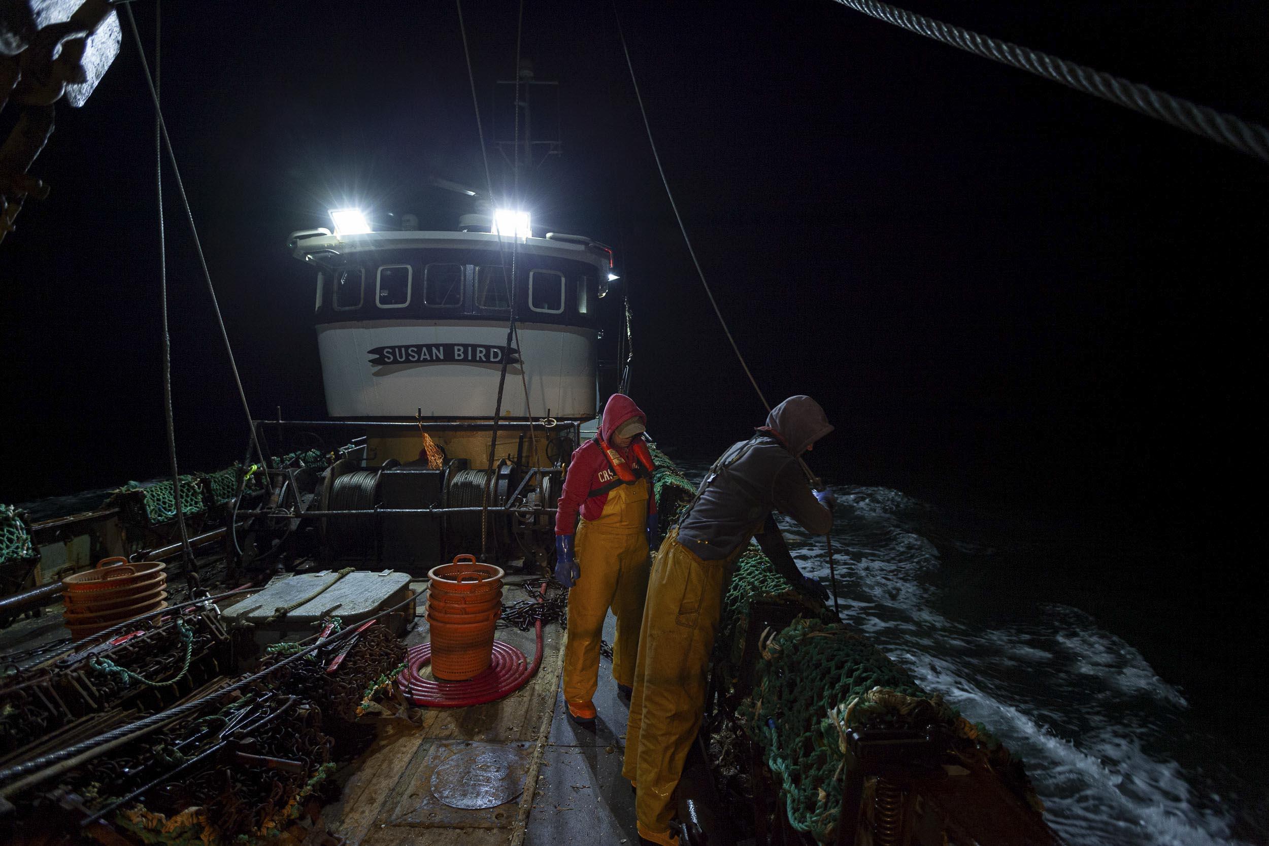 The Susan Bird fishing trawler on the sea at night. Two fishermen are looking over the side of the boat at the scallop dredger.