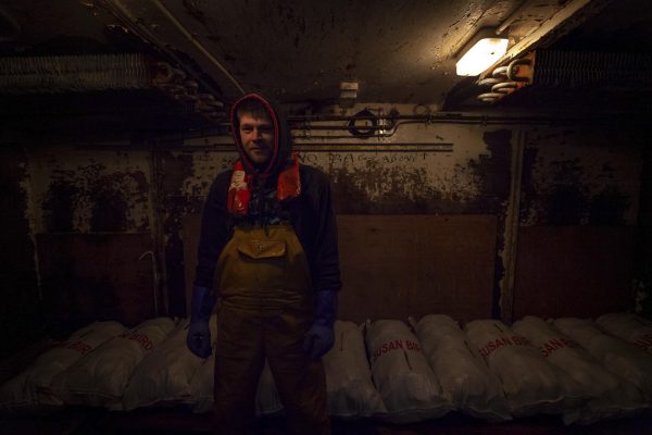 A fisherman standing inside the cold room storage of a fishing vessel surrounded by bags of scallops. The light is dim.