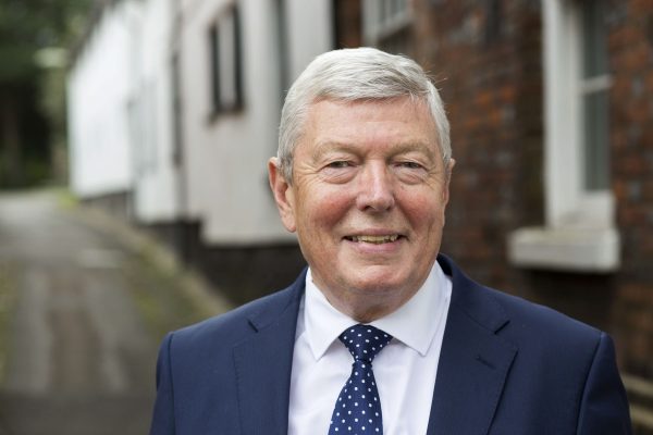 British politician Alan Johnson stands smiling in a street. He wears a blue suit and tie.