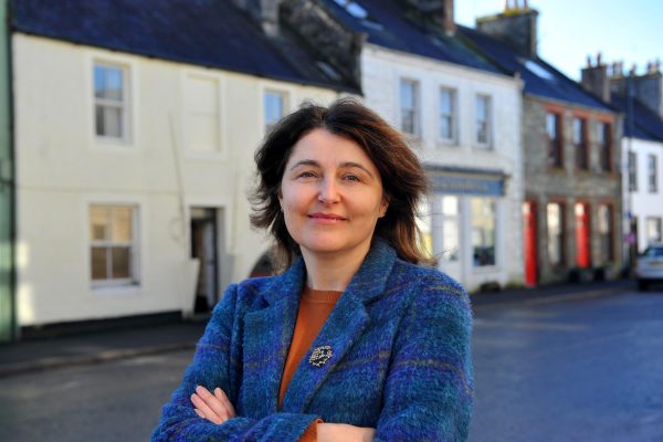 Karen Campbell stands in Wigtown, Sccotland's National Book Town. Her hands are folded she is smiling. Terraced houses behind her.