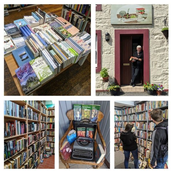 Well Read Books. Wigtown. Proprietor Ruth Anderson standing in the doorway to the shop, above her the shop sign. Inside people are browsing the books, a typewriter sits on a chair surrounded by books.