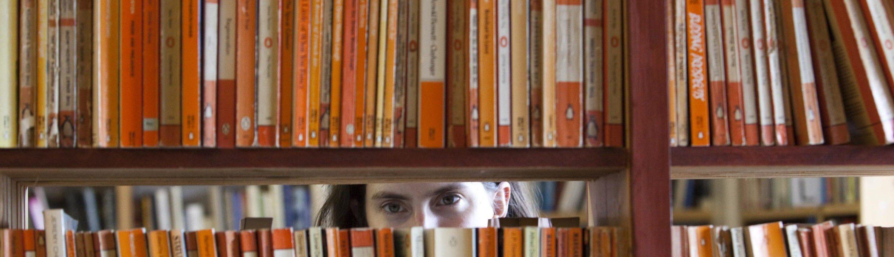 Shelves of old paperback books, a person is peering through the gap in between the top and bottom shelves.