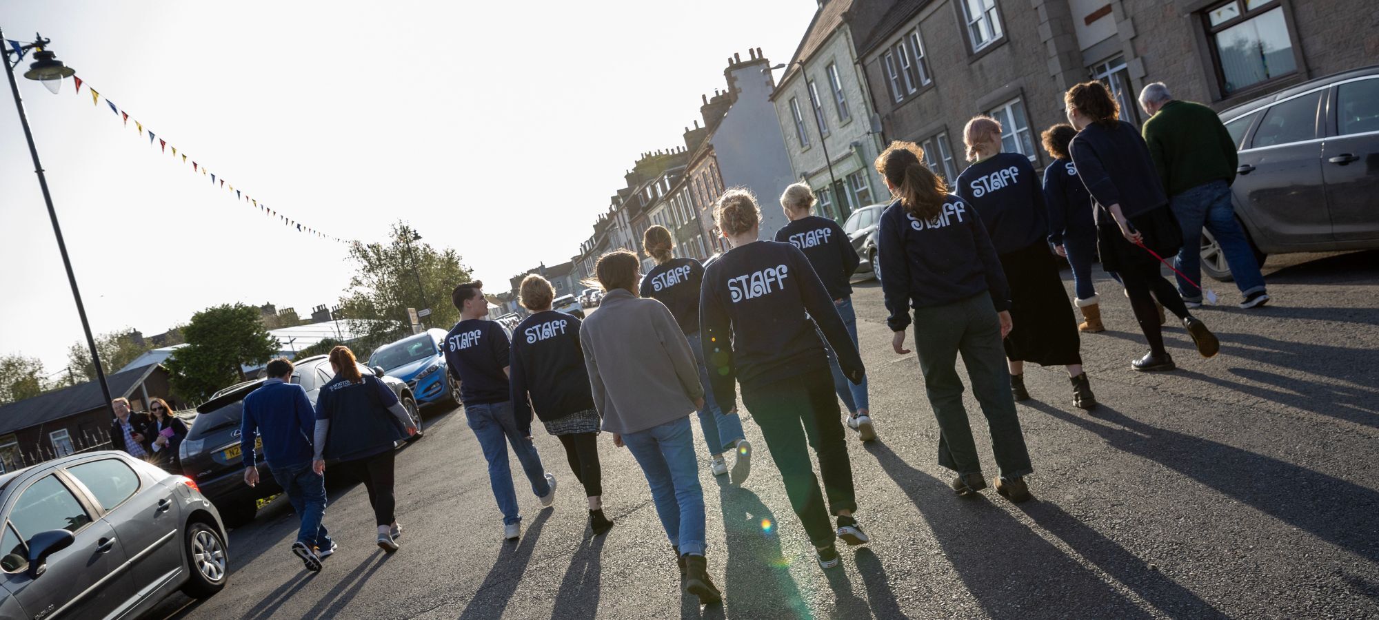 13 people, many wearing Wigtown Book Festival sweatshirts with 'Staff' written on the back, walk away from the camera. Low sunlight casts long shadows. They are walking down North Main street in Wigtown, with parked cars, building and bunting in the background.