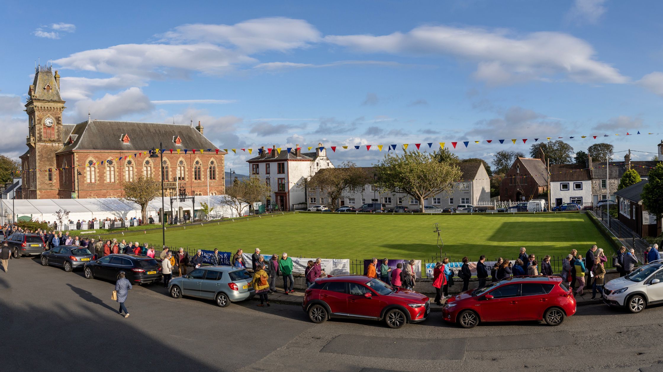 Panorama image of Wigtown with a large queue of people around the Bowling Green, at the centre of town. The County Buildings is in the background, with colourful bunting hung between lampposts.