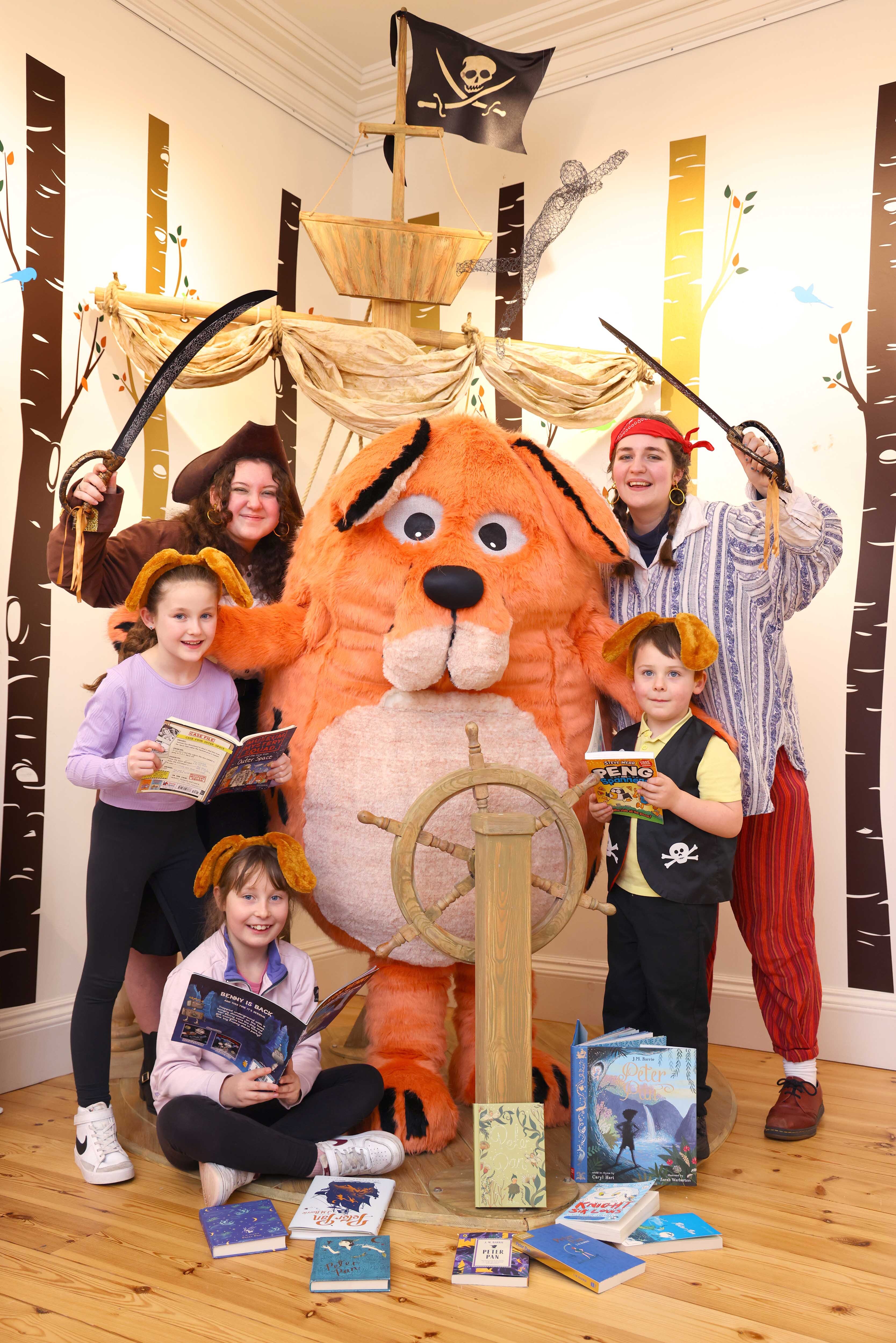 The Big DoG mascot is surrounded by children wearing matching orange dog ears and the Moat Brae Storyweavers dressed as pirates. The are gathering around a pirate ship play display with festival titles.