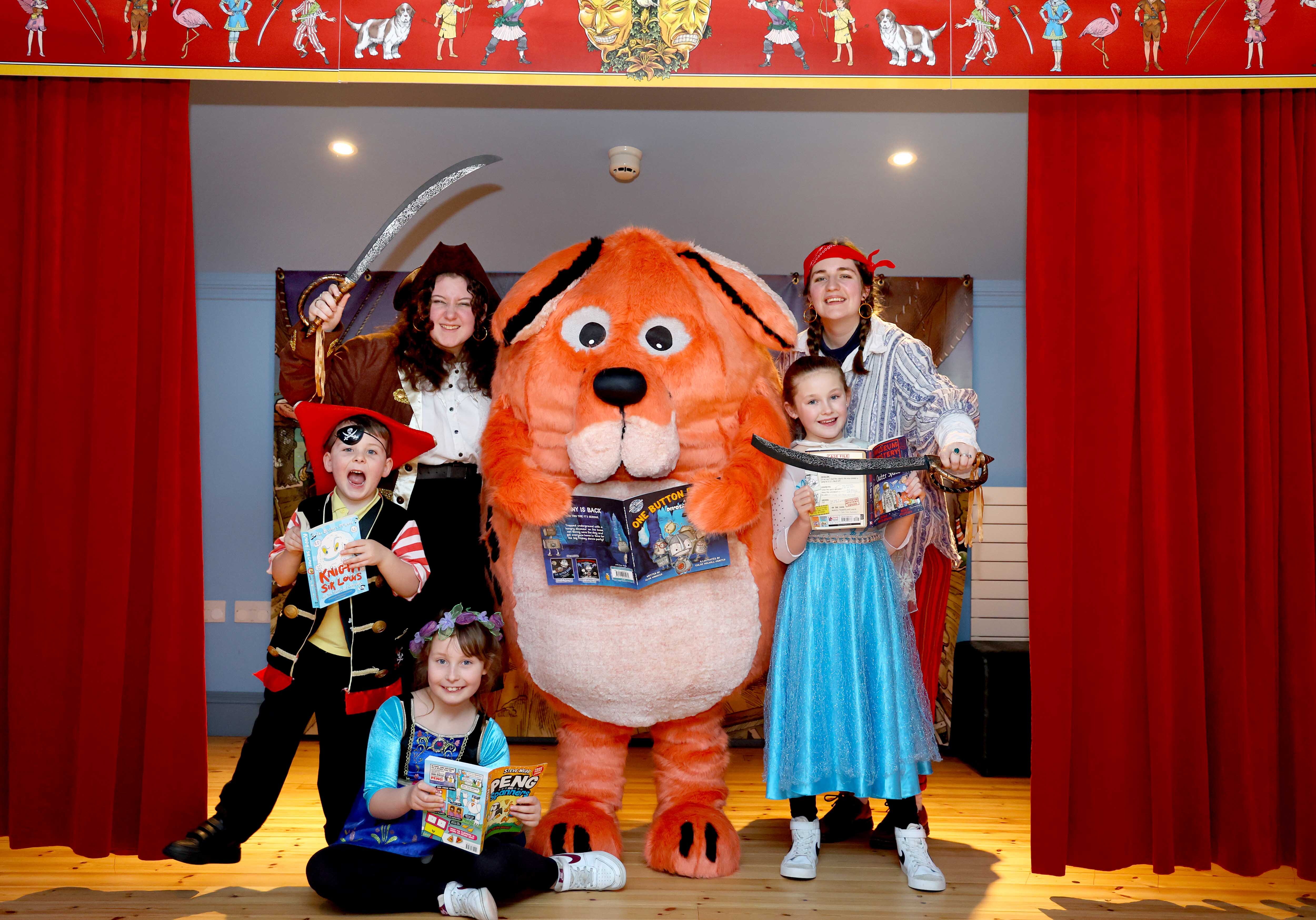 The Big DoG mascot is surrounded by children wearing matching orange dog ears and the Moat Brae Storyweavers dressed as pirates. They are stood in the children's theatre at Moat Brae.