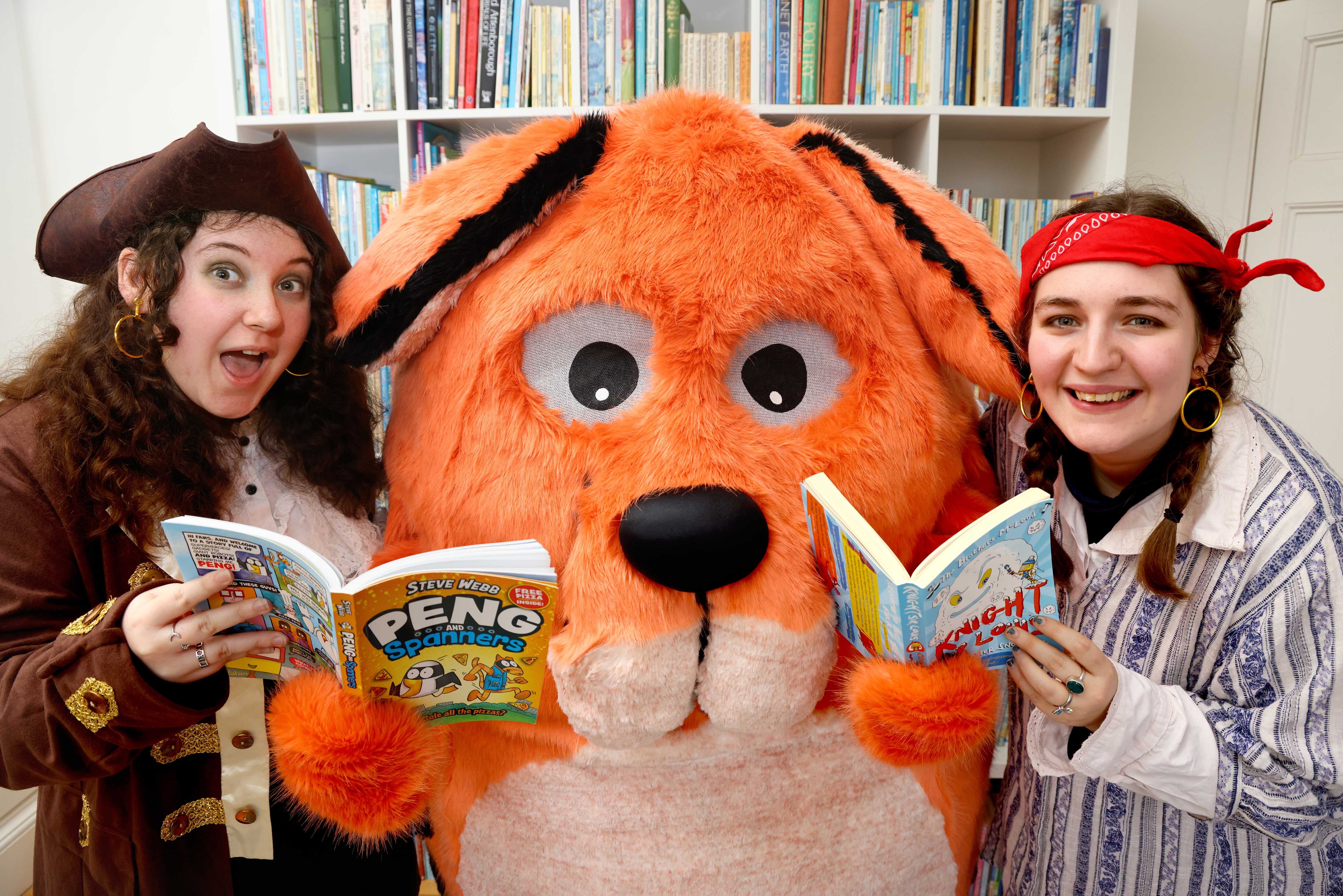 Big DoG and the Moat Brae Storyweavers, dressed as pirate, are stood in front of a book case, holding Big DoG festival titles.