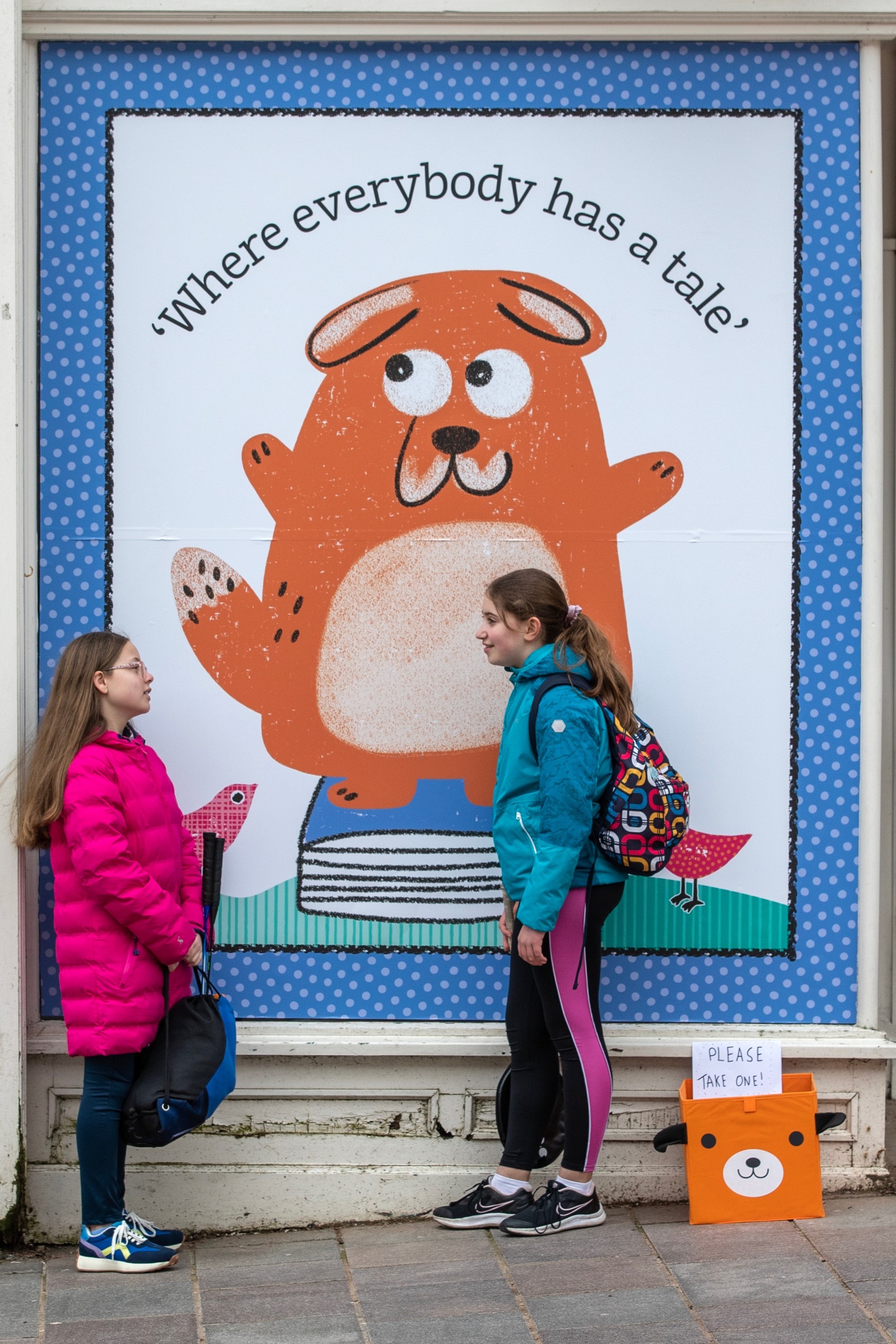 Two children stand talking outside Big DoG HQ in front of a large window decoupage featuring the character Big DoG, an orange dog, with the caption 'Where everybody has a tale' above.