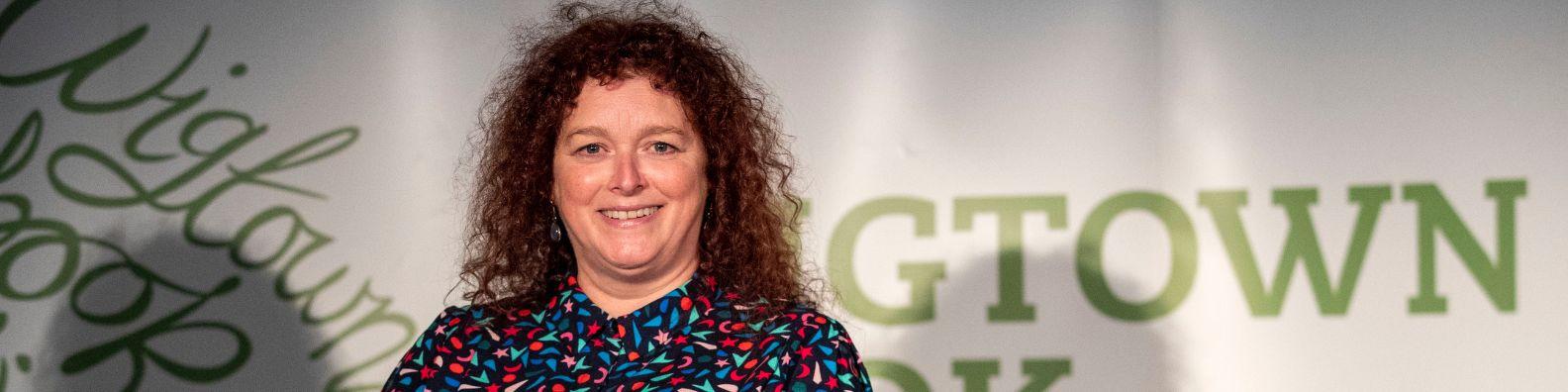 gtown Book Festival Poetry Prize winner 2022 Julie Laing. She stands in front of the green and white Wigtown Book Festival logo.