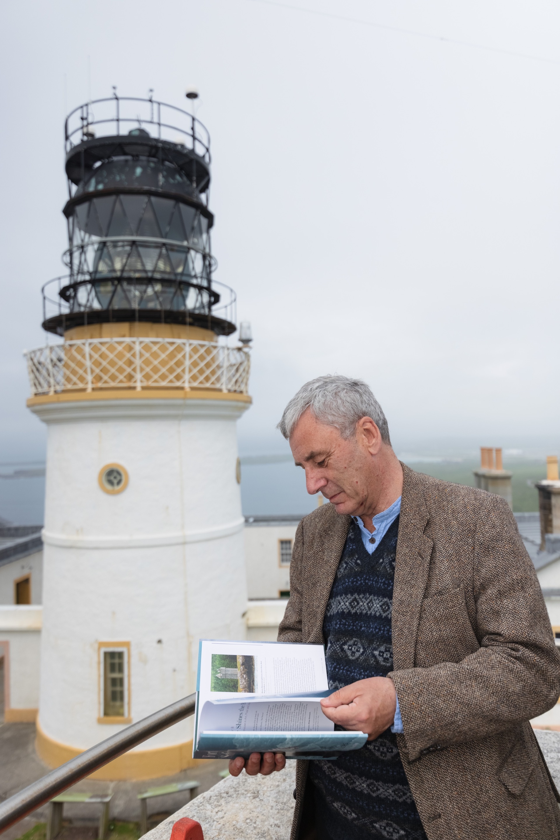 Poet Donald S Murray stands reading a book, in front of a lighthouse.