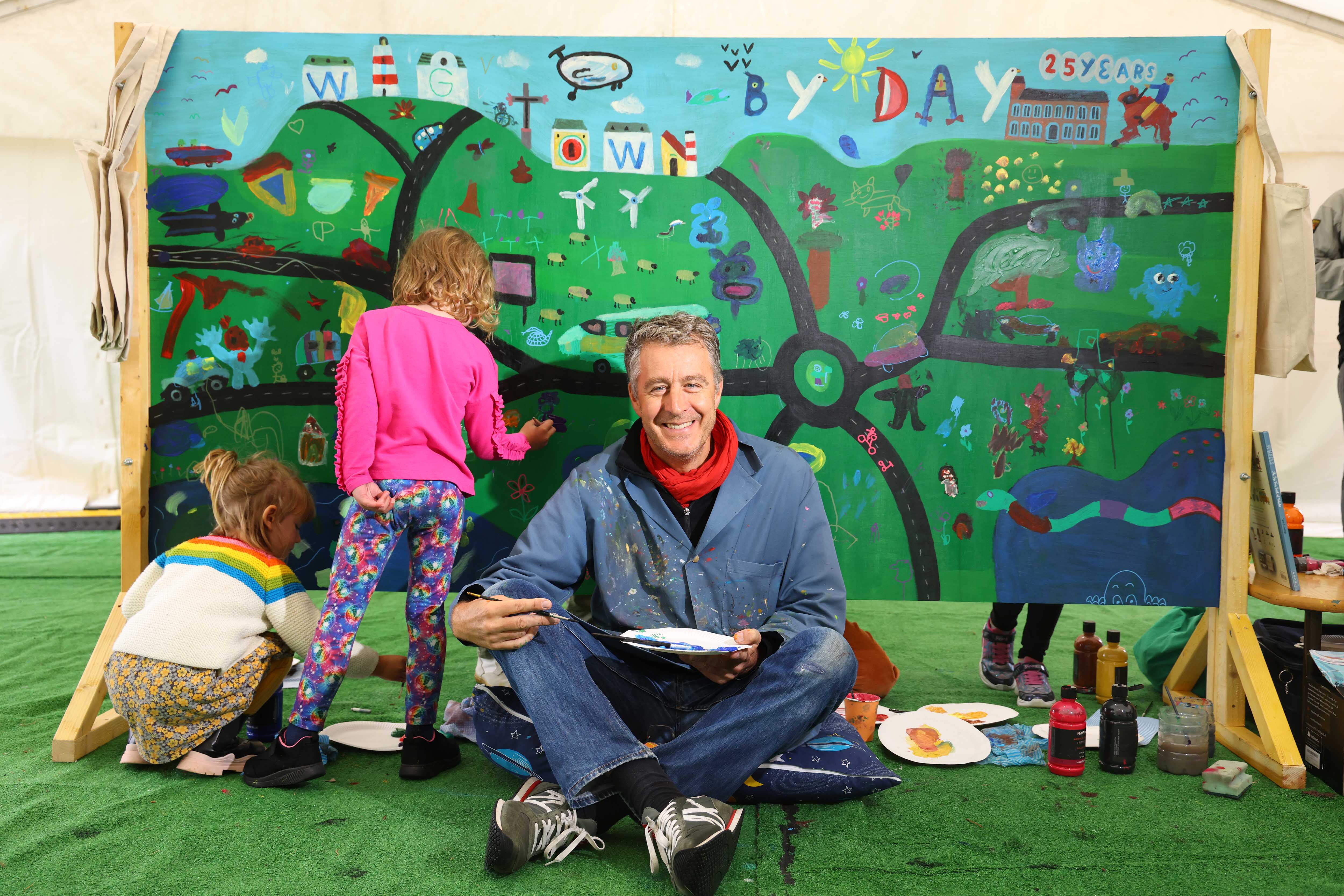 Artist Tom Schamp in front of a collaborative artwork made with the children of Wigtown. Two children are stood behind him painting.