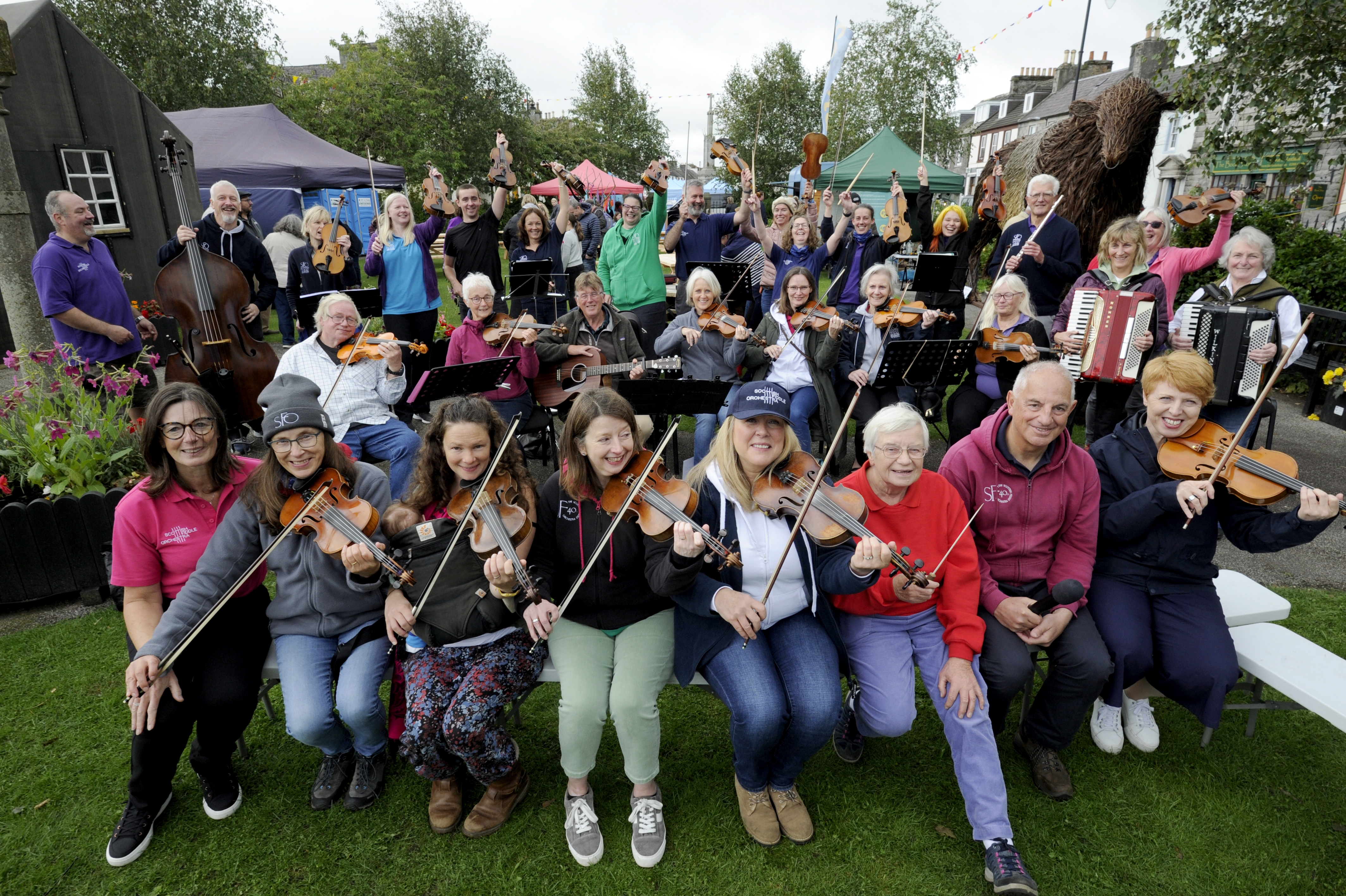 Many people sitting in the Wigtown gardens playing fiddles for the Fiddlers Fair outdoor event at the Wigtown Book Festival. . A few have guitars and accordions, another has a double bass. The players in the background are raising their instruments in a cheer.