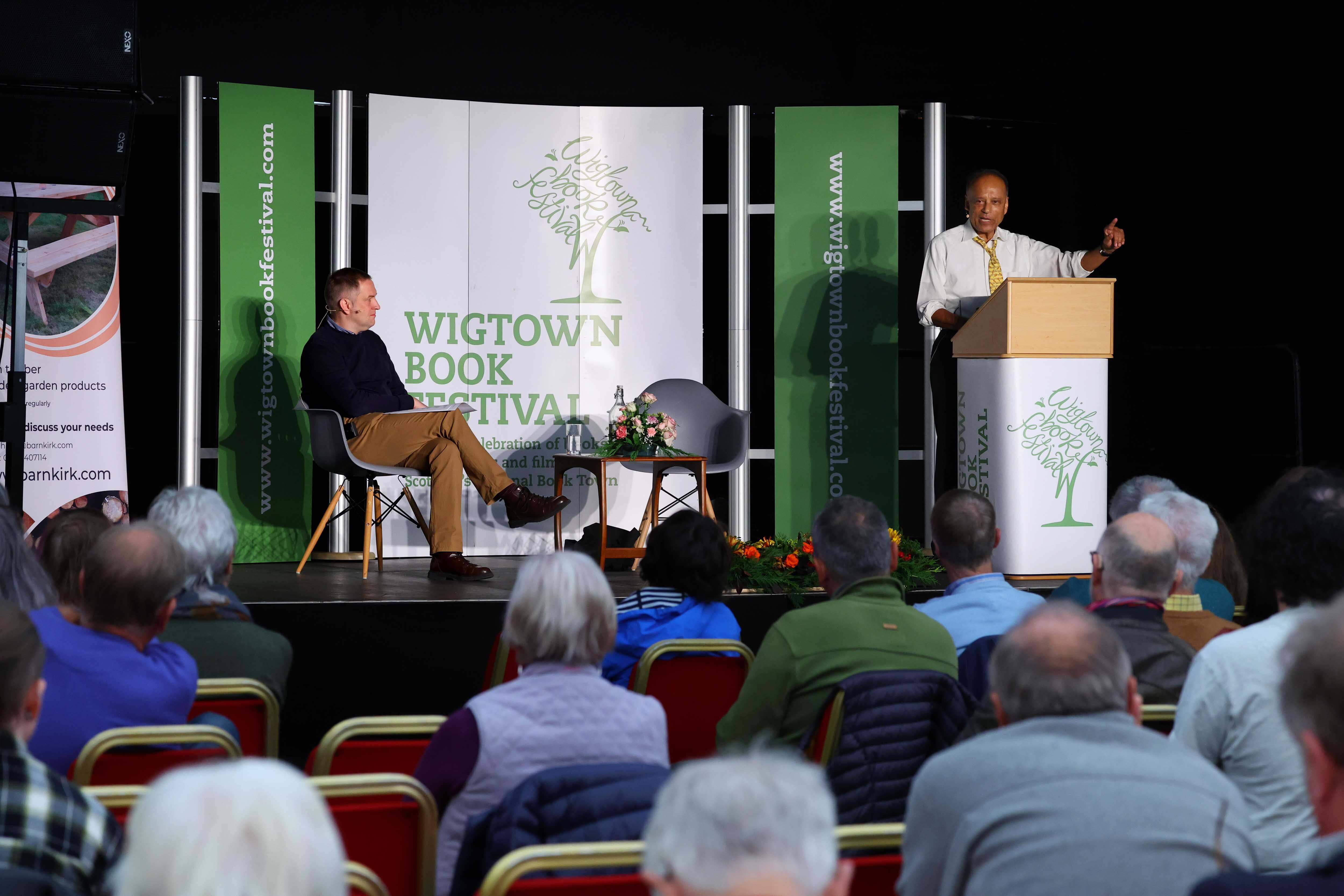 Professor Sir Partha Dasgupta speaks from behind a lectern on stage at Wigtown Book Festival. Chair Graeme Stewart sits in a chair across the stage in front of a Wigtown Book Festival Banner. Audience members are in view in the foreground.