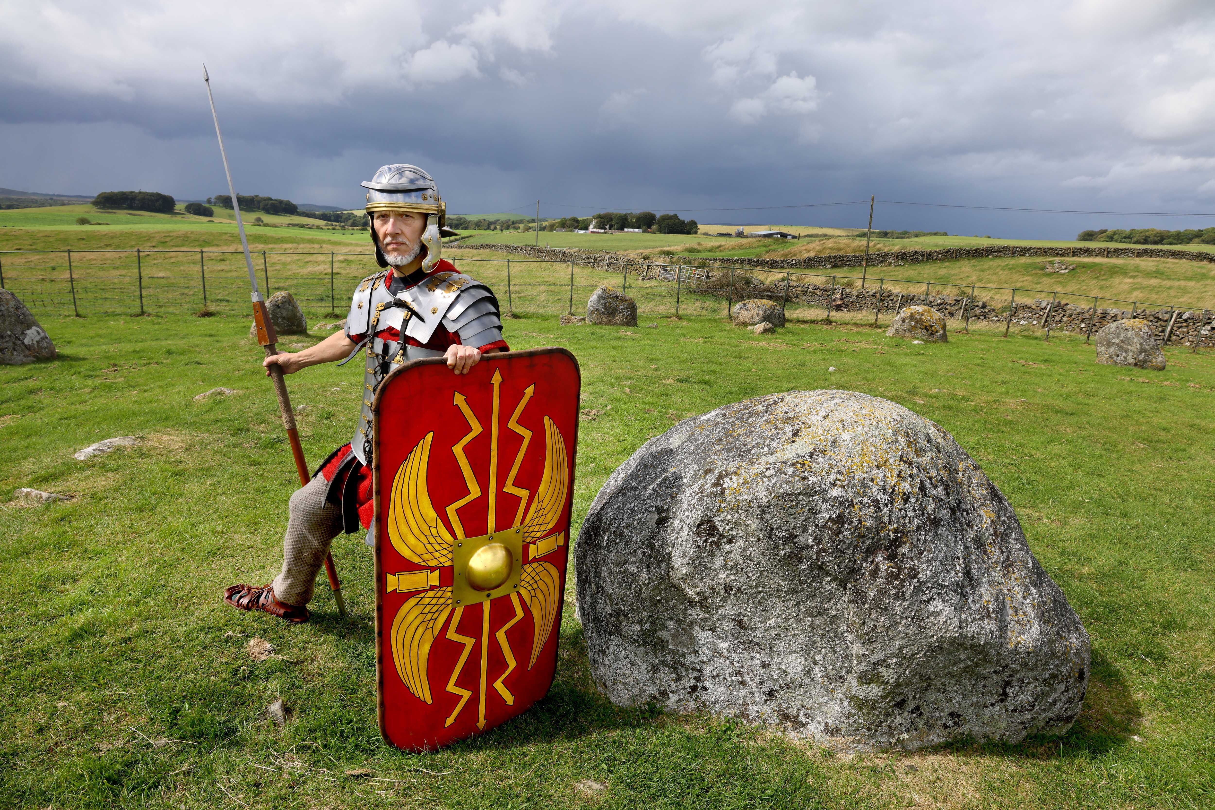 Matthew Shelley dressed as a Roman centurion, stood next to a boulder on a hill, looking stoic.