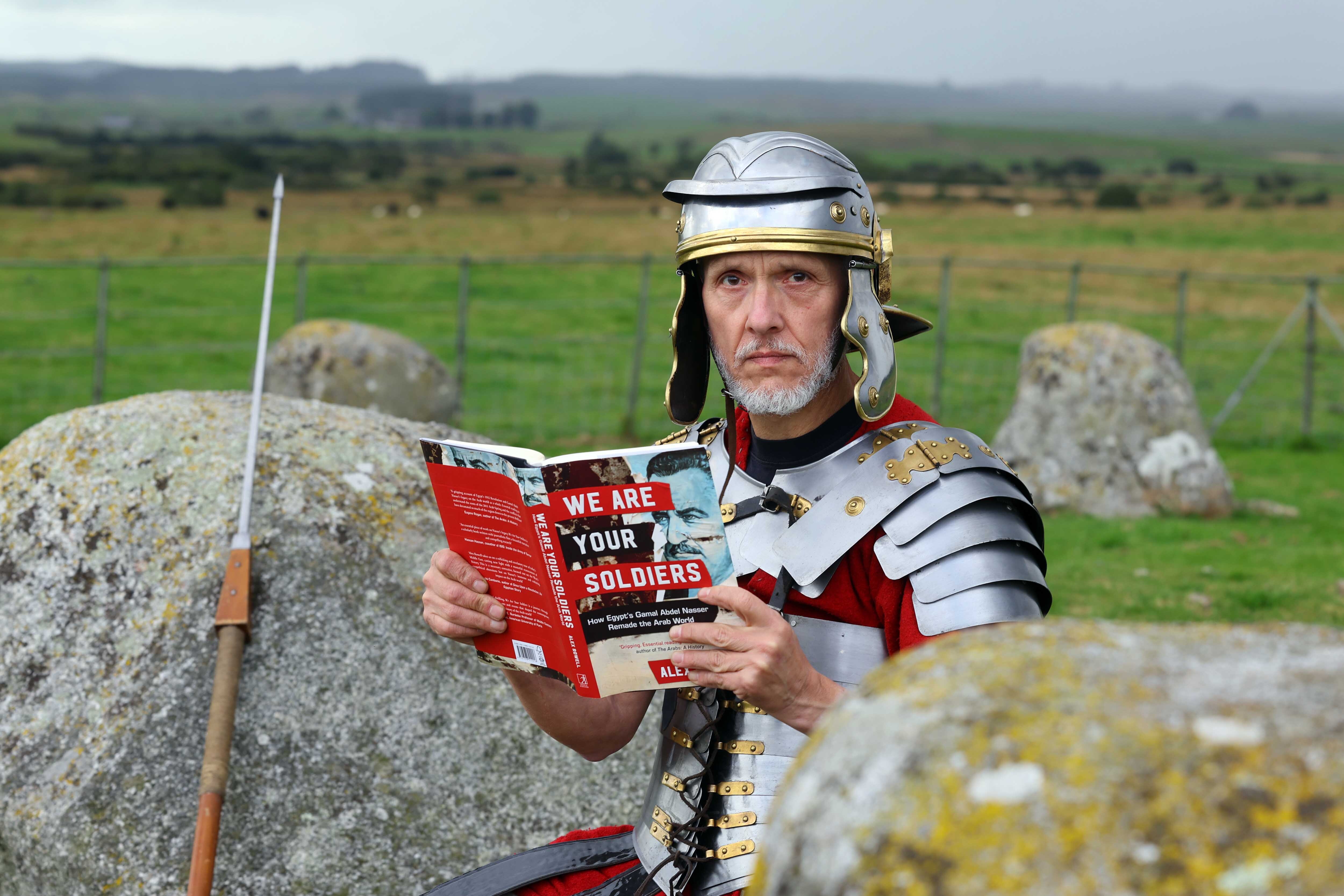 Matthew Shelley dressed as a Roman centurion, sits on a boulder, his spear resting nearby, reading a 2024 Wigtown Book Festival title and staring into the camera.