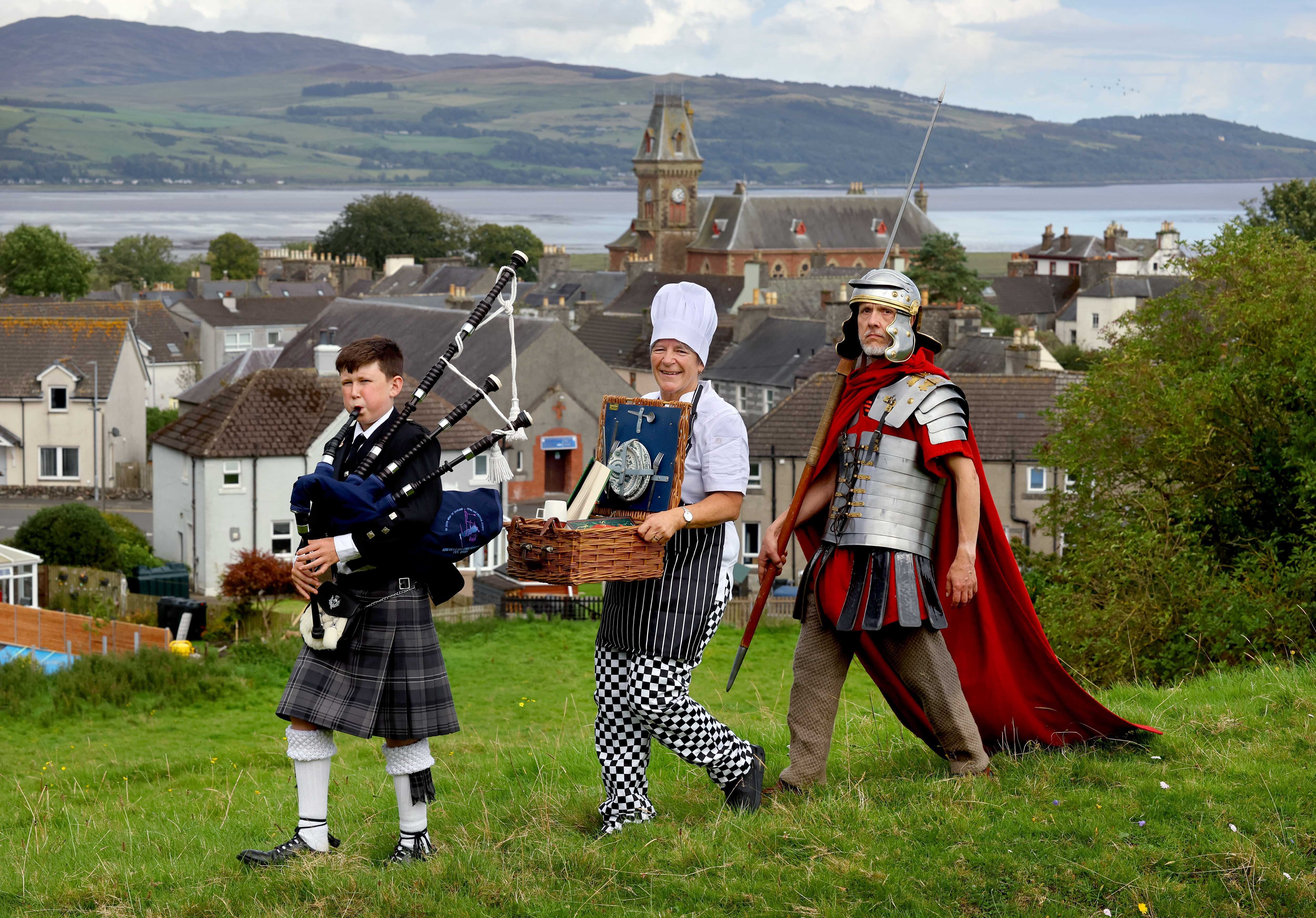 One piper piping, one chef carrying a picnic basket and one centurion marching, on a hill with Wigtown and Wigtown bay behind them.