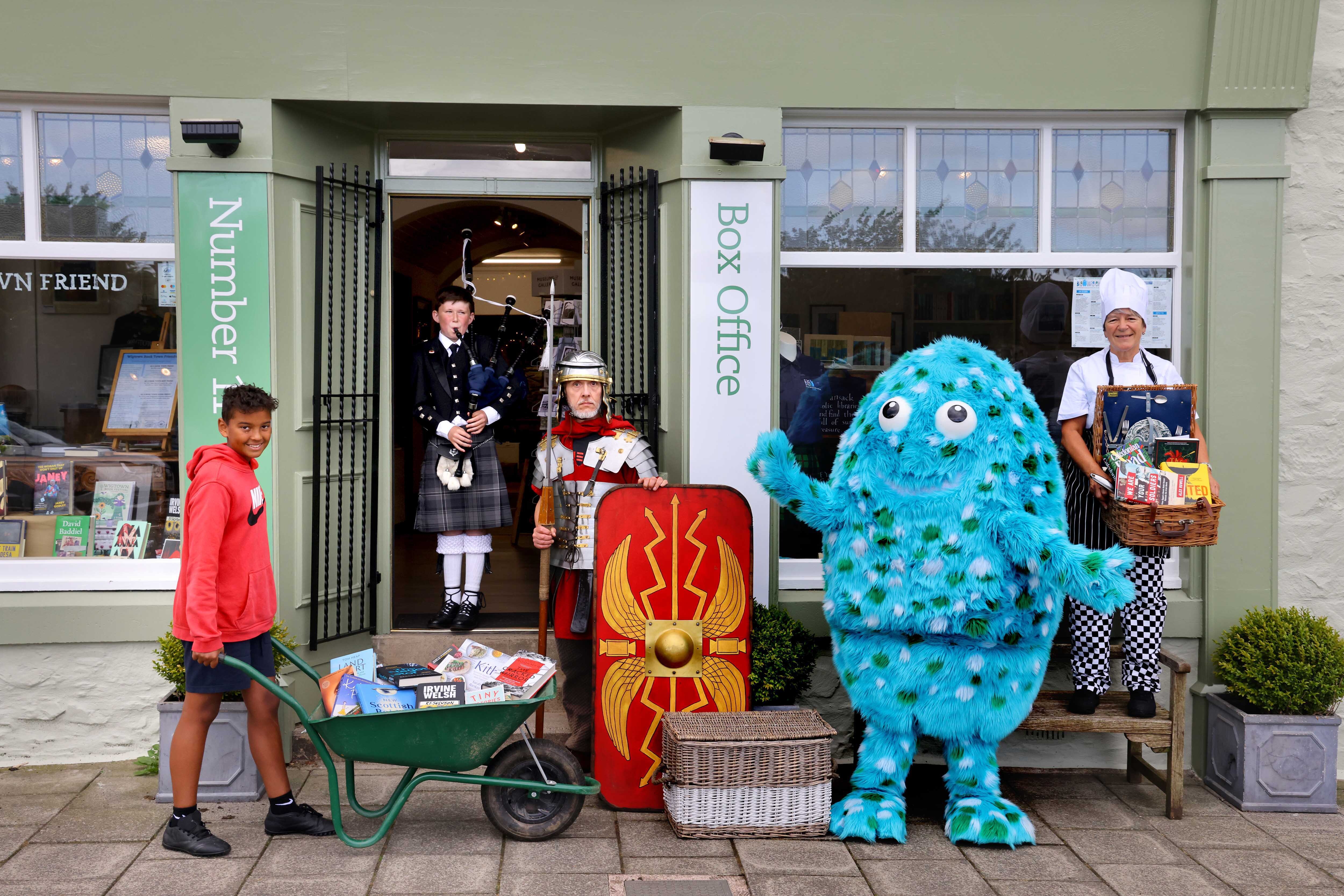 A chef, a roman centurion, a bagpiper and a young person pushing a wheelbarrow filled with books stand outside Number 11 Bookshop