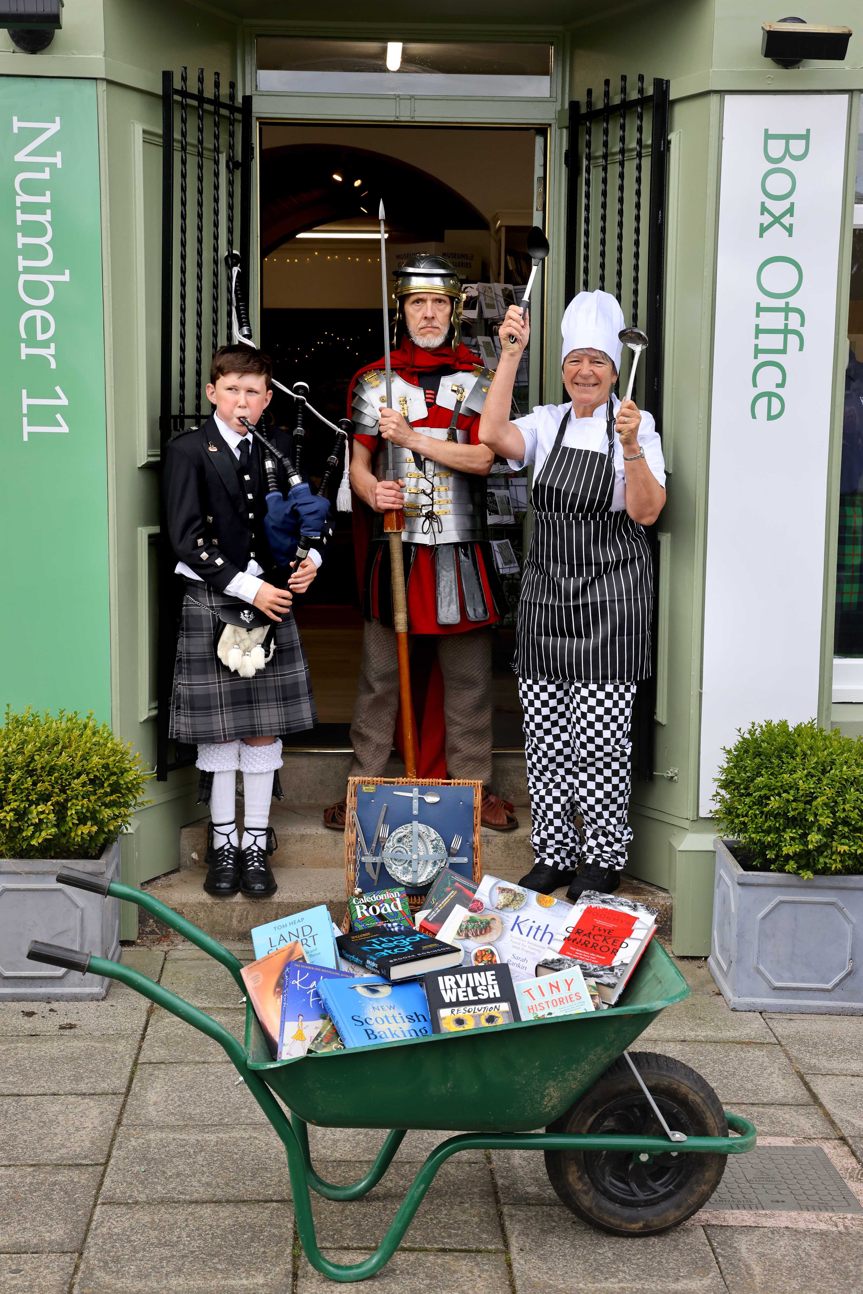 A chef, a roman centurion, a bagpiper and a wheelbarrow filled with books outside Number 11 Bookshop, portrait.