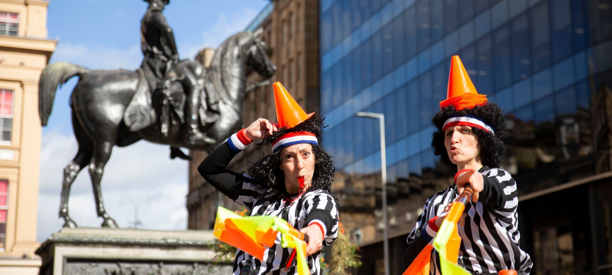 The Refs, two children's street entertainers, are stood in a city square, wearing black-and-white-striped referee t-shirts, small orange cones on their heads and holding out two orange flags. They are both wearing animated expressions, one person is blowing a red whistle.