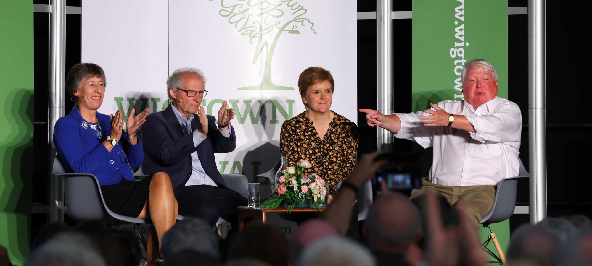 Left to right Liz Smith, Henry McLeish, Nicola Sturgeon and Brian Taylor, seated on stage in front of Wigtown Book Festival banner. Audience blurred in the foreground.