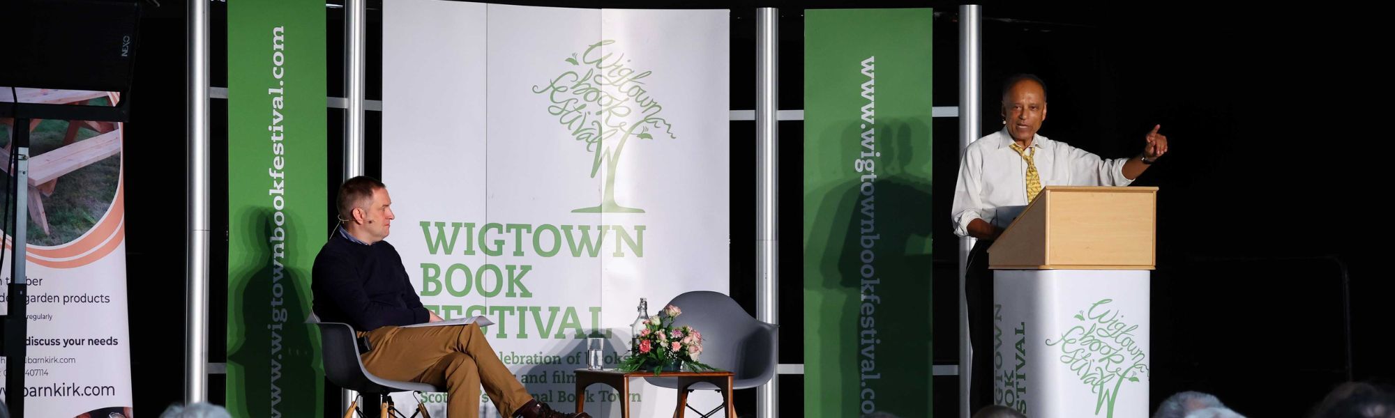 Professor Sir Partha Dasgupta stands on stage at Wigtown Book Festival, speaking behind a lecturn. Chair Graeme Stewart sits at a table nearby. They are both in front of Wigtown Banners.