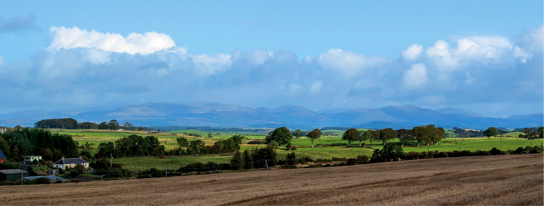 Rural view of the Galloway Hills. A ploughed field with green fields and the hills in the distance. Trees and hedgerows with a few houses. Cumulus clouds in a blue sky.