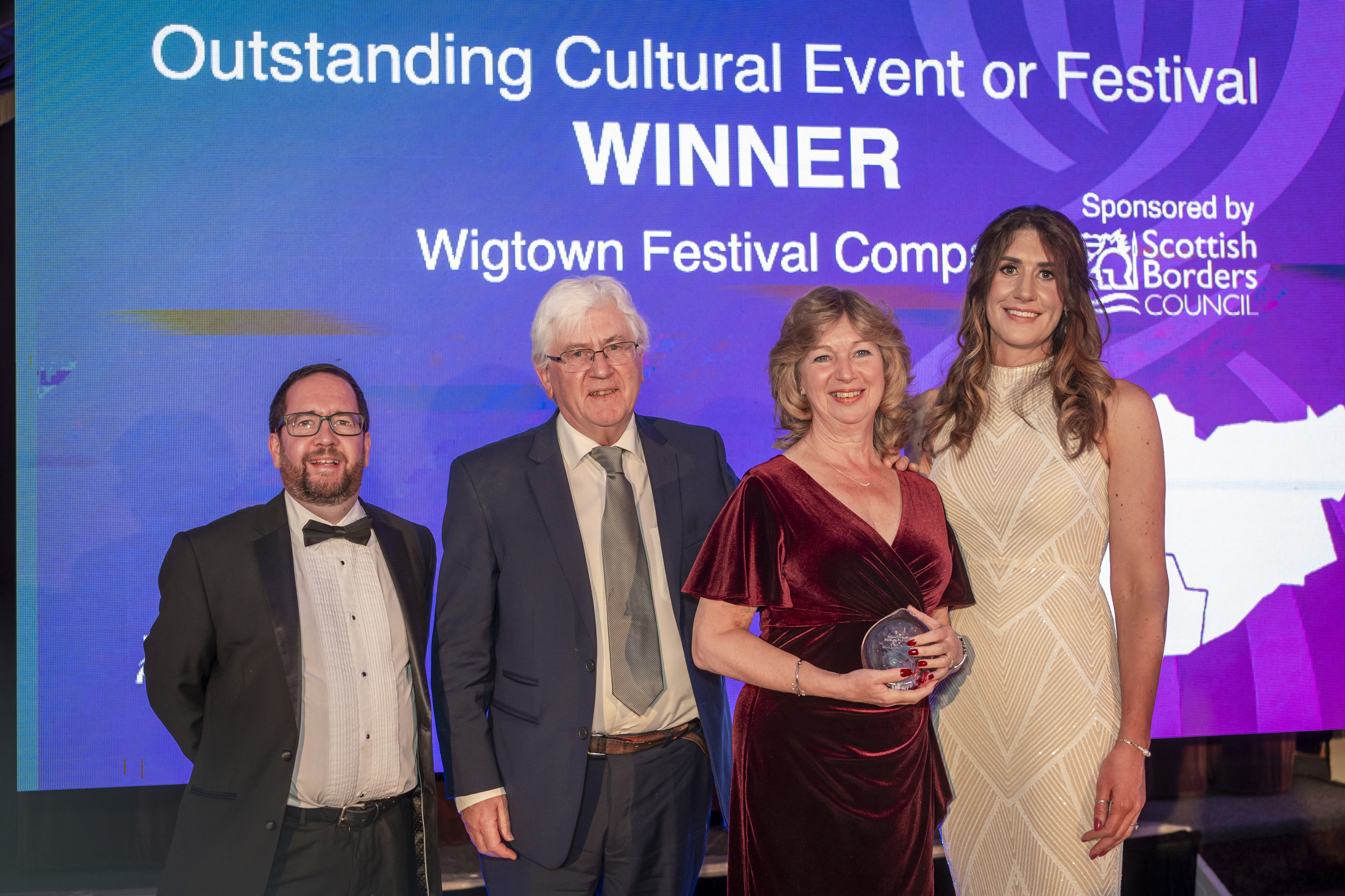 South of Scotland Thistle Award ceremonies, featuring hosts and Wigtown board members on stage holding the award. A screen behind them reads 'Outstanding Cultural Event or Festival Winner, Wigtown Festival Company', in white text against a blue and purple background.