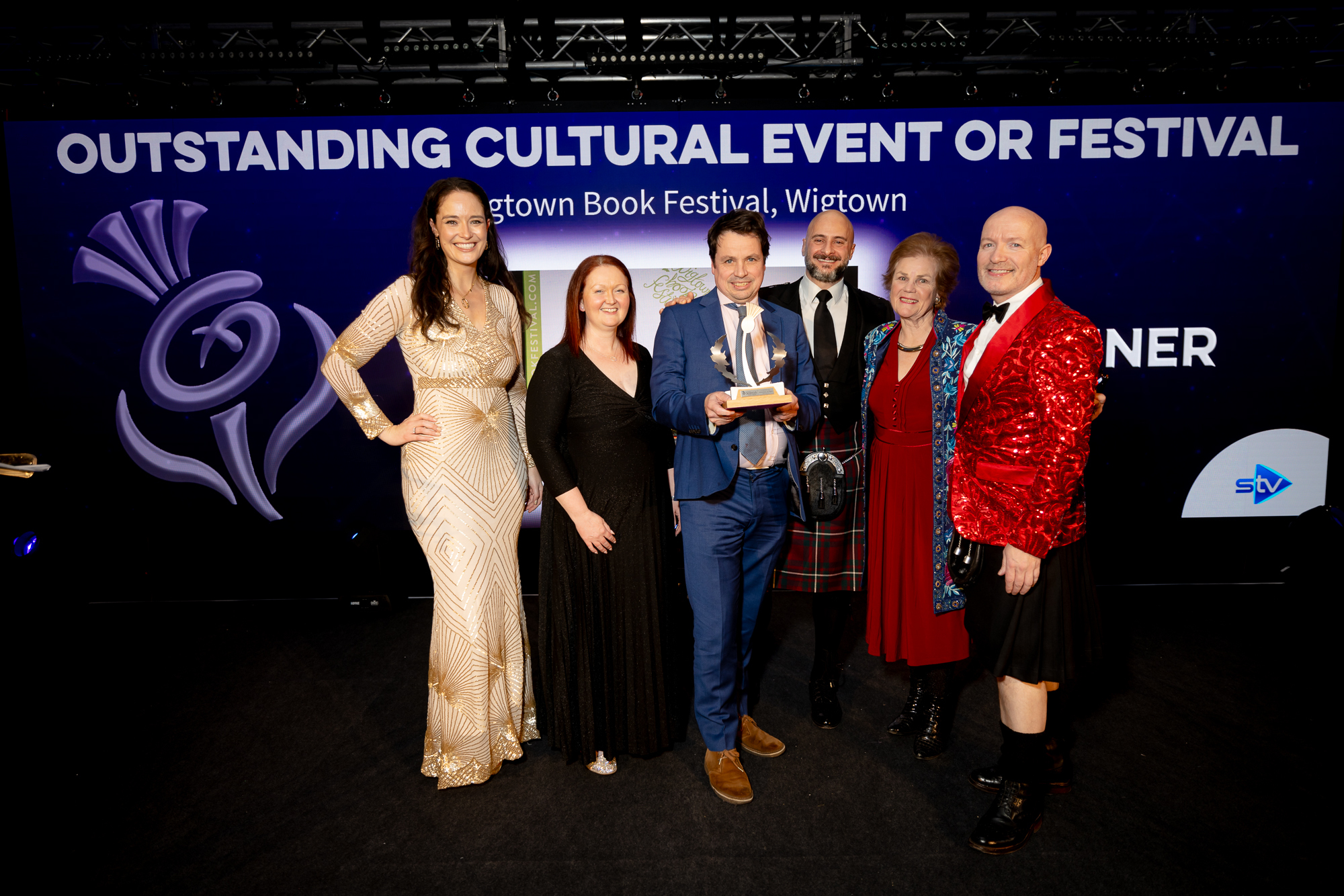 Six people on stage, including Operational Director Anne Barclay, Artistic Director Adrian Turpin holding an award, Chair of Wigtown Festival Company Cathy Agnew and three Scottish Thistle Award presenters.