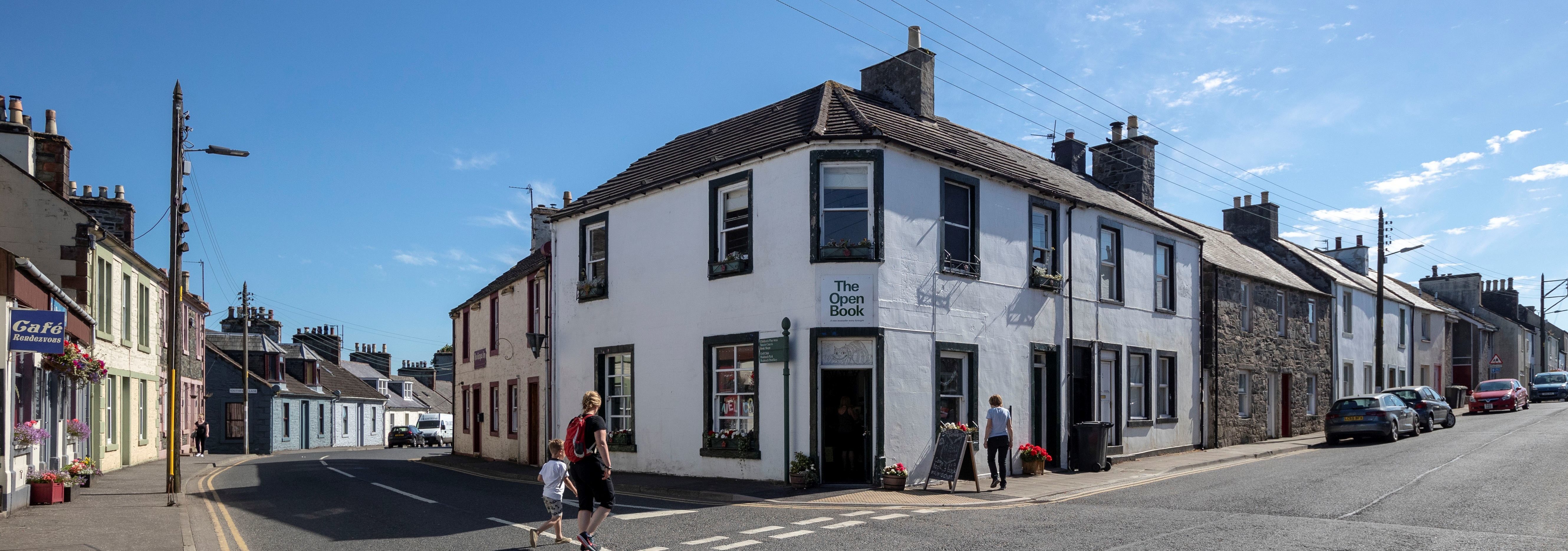 Corner view of the Open Book Bookshop in Wigtown. An adult and child are crossing the road, cars parked in the street, a telegraph pole on the left.