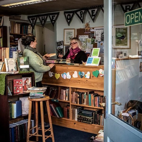 Through the front door into The Open Book bookshop. A person stands behind the counter serving. Many books are sitting on shelves, a small stool has more stacked books on it.