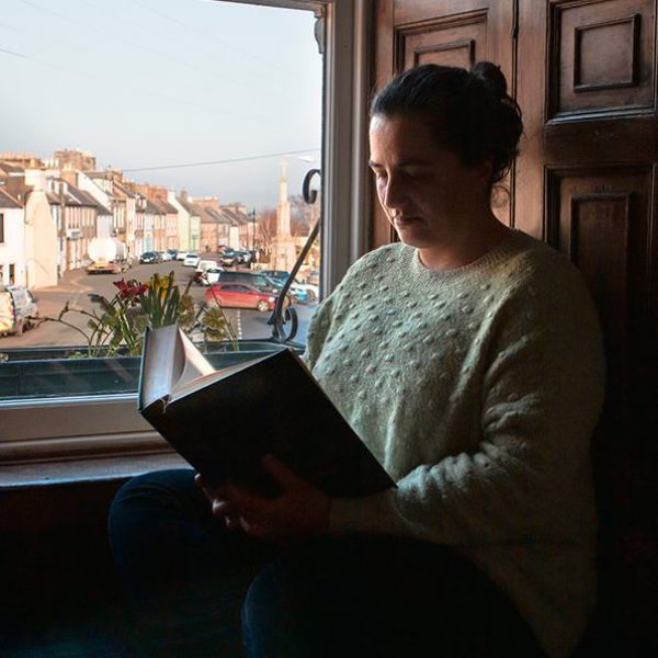A person sits in front of a large sash window reading a book. Behind them through the window is Wigtown Main Street with parked cars. houses and the Mercat Cross.