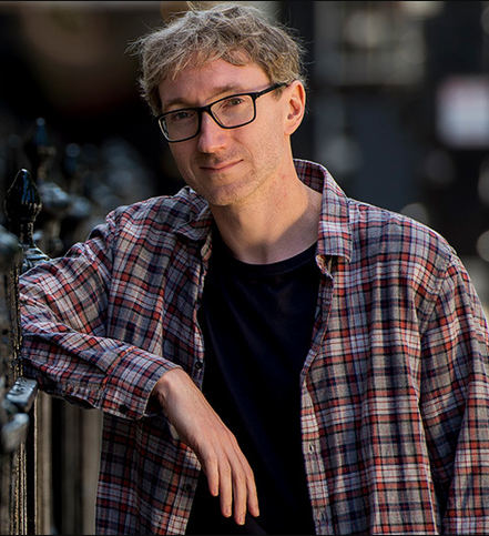 Stuart Turton is standing resting his arms on black railings.