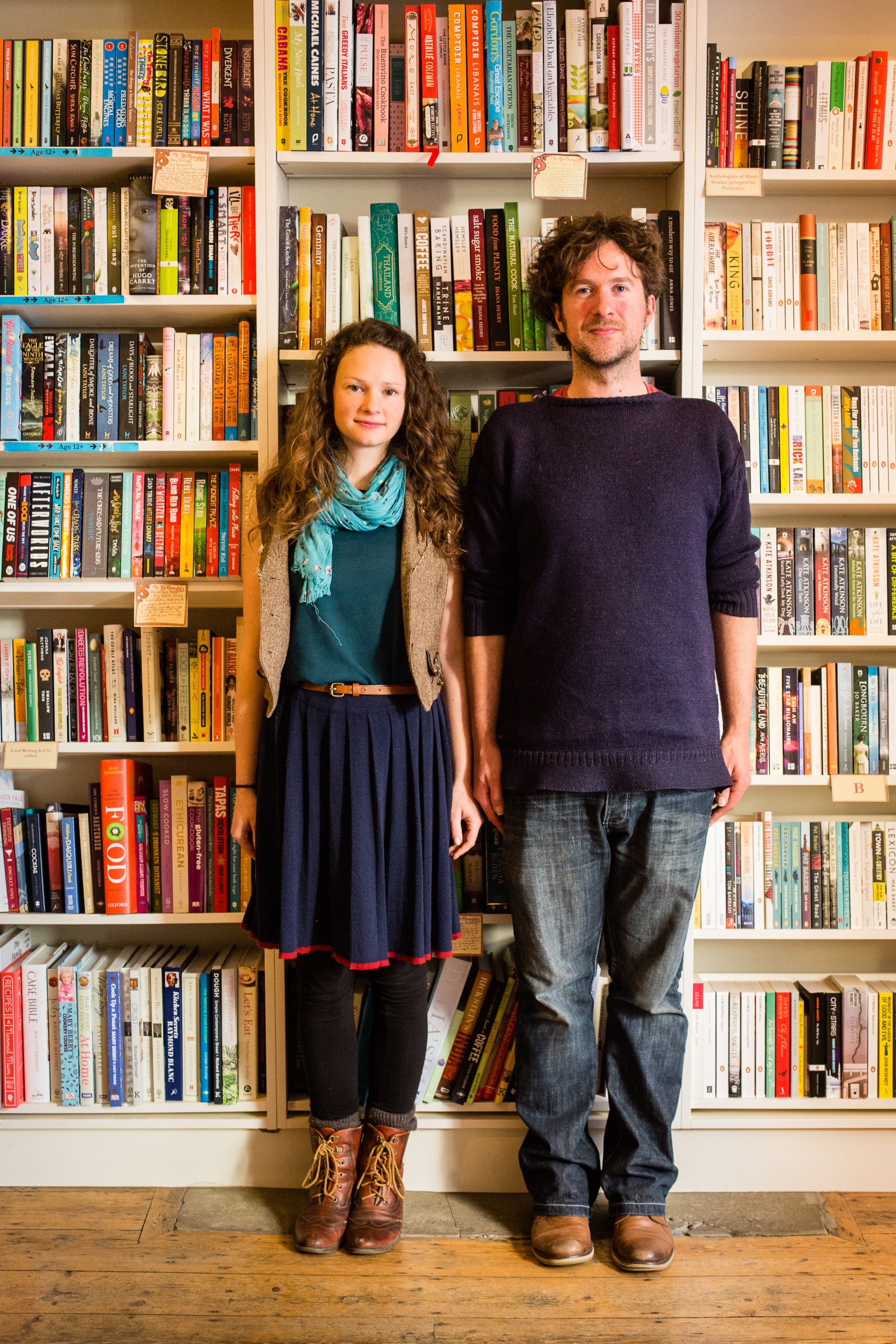 Ben Please and Beth Porter of the Bookshop Band stand in front of a wall of bookcases.