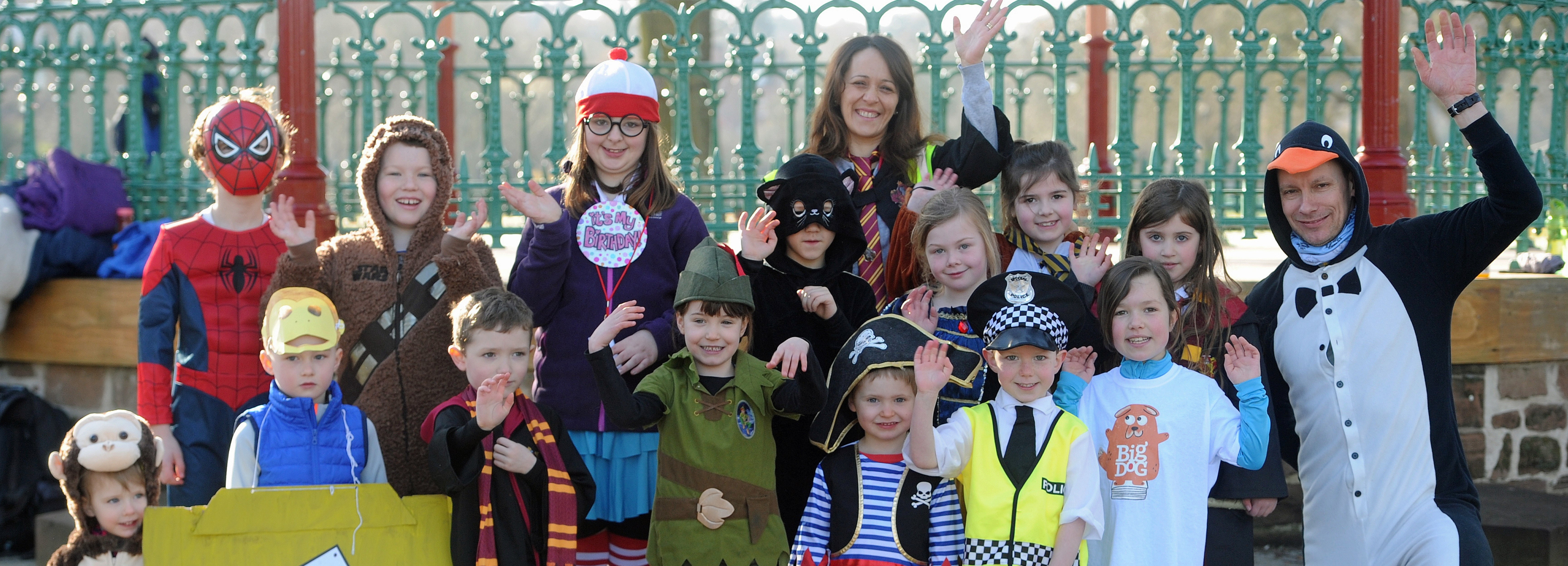 A group of children dressed up as various book characters stand waving and smiling at a Children's Book Festival event. Green railings are behind them.