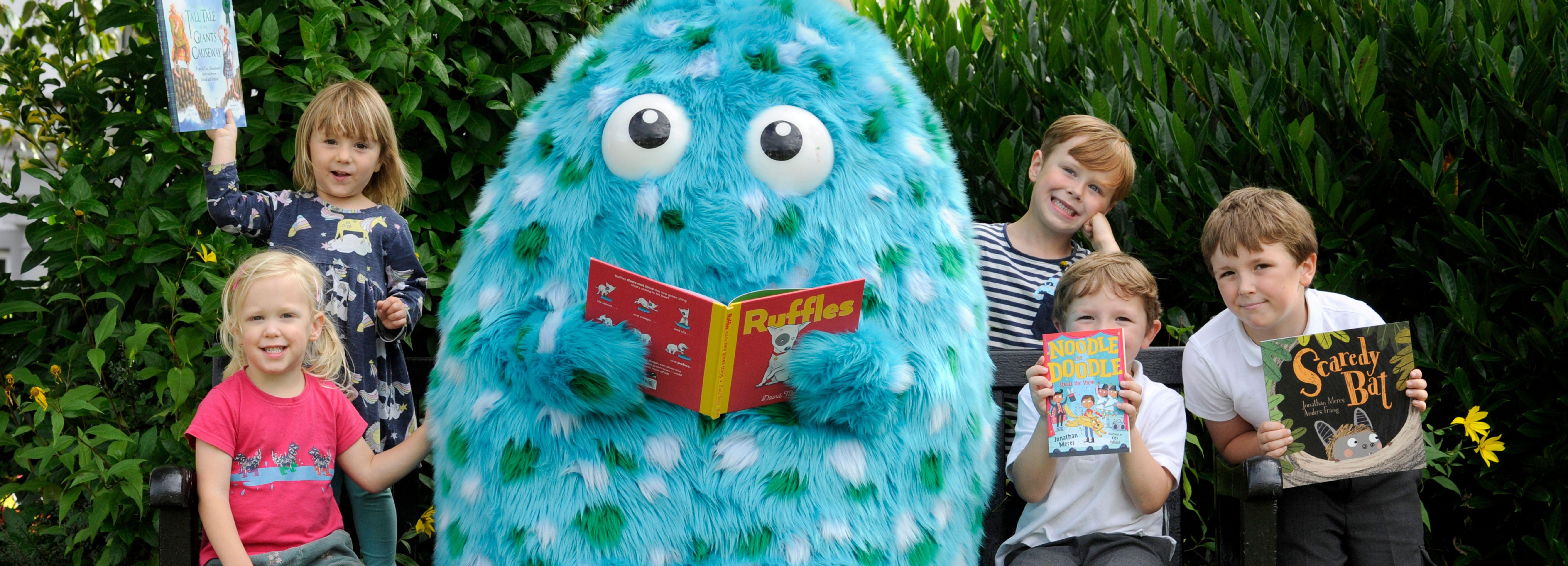 Bigwig, our children's book festival mascot, stands with various children in Wigtown gardens reading a book, The children are smiling and holding books up.