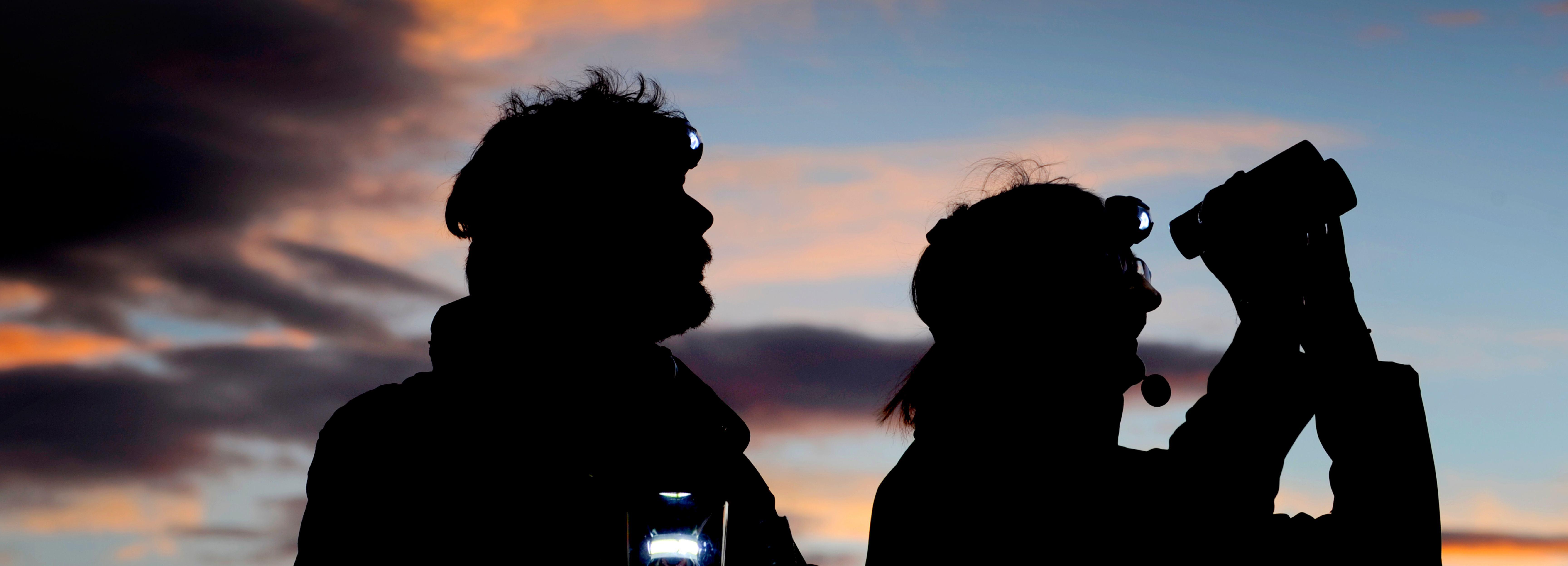 Dan Richards and Elizabeth Tindal in silhouette against a sunset sky. Elizabeth is holding up binoculars.