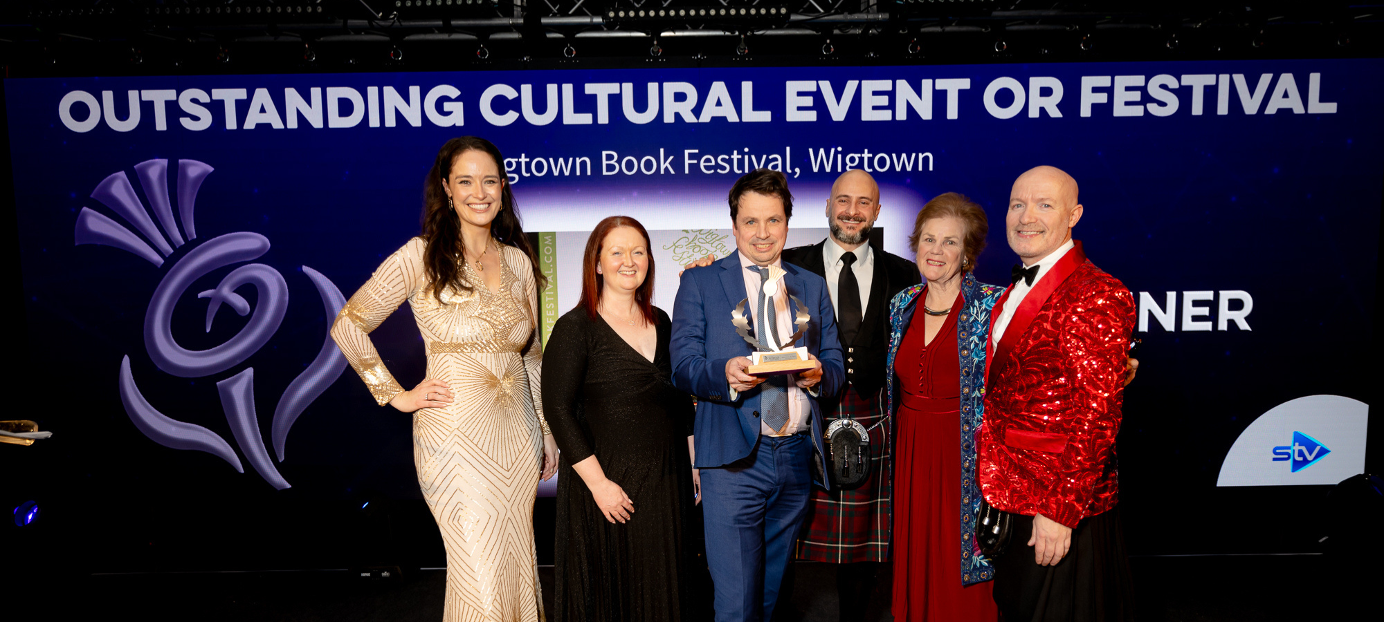 Six people on stage, including Operational Director Anne Barclay, Artistic Director Adrian Turpin holding an award, Chair of Wigtown Festival Company Cathy Agnew and three Scottish Thistle Award presenters.