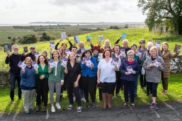 A crowd of staff and volunteers stand in front of a view over the Wigtown marshes. Each of them is holding a 2023 programme brochure.