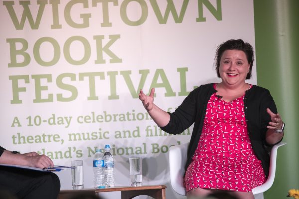 A lady sitting in a chair upon a stage wearing a bright pink dress, talking at an event at Wigtown Book Festival . A table containing bottles of water beside her.