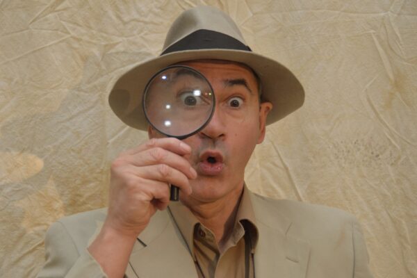 Clydebuilt Puppet theatre Dinosaur Detective image, portrait of a person wearing a beige suit and hat, holding a magnifying glass to their eye.
