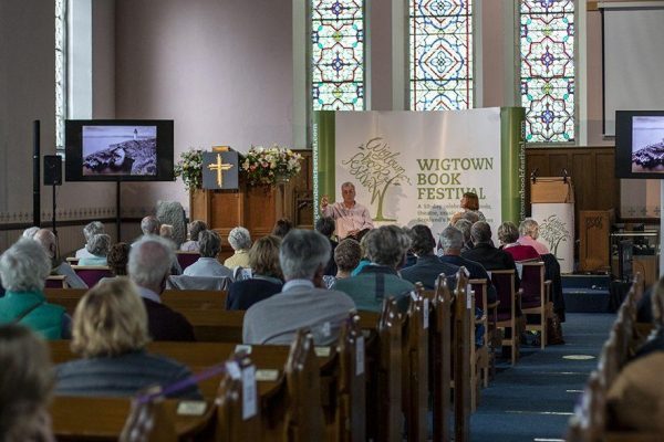 A Wigtown Book Festival event taking place in a church. The audience are sitting in the pews listening to an author talking on stage. Two screens are showing slideshows.