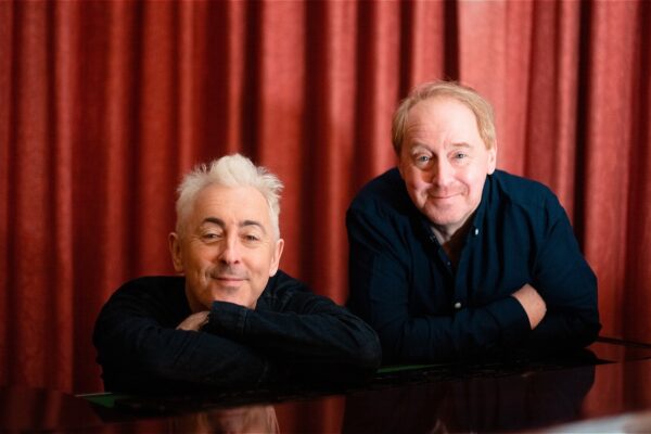 Alan Cumming and Forbes Masson leaning on a piano in front of a stage curtain.