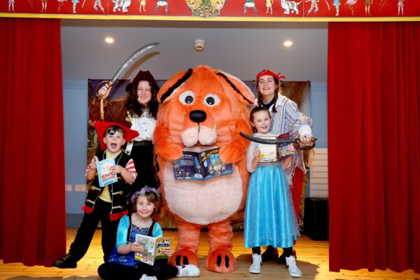 The Big DoG mascot is surrounded by children wearing matching orange dog ears and the Moat Brae Storyweavers dressed as pirates. They are stood in the children's theatre at Moat Brae.