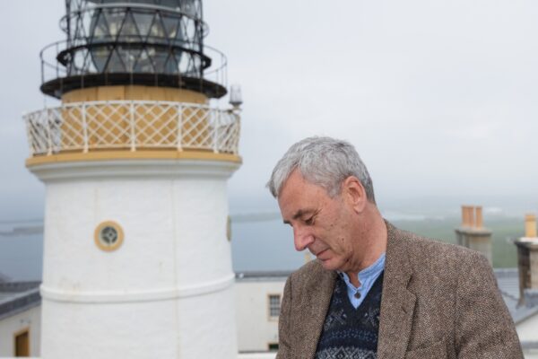 Poet Donald S Murray stands reading a book, in front of a lighthouse.