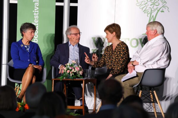 Left to Right: Liz Smith, Henry McLeish, Nicola Sturgeon and Brian Taylor, seated on stage at Wigtown Book Festival, around a table with flowers on. Audience blurred in foreground.