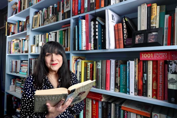 Lee Randall stands in a bookshop smiling and holding open a book. The bookshelves behind her are full of books.