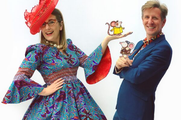 Author-illustrator Sarah McIntyre stood on the left, wearing a blue and red patterned dress with flared sleeves and a large red hat. Author-illustrator Philip Reeve stood to the right, wearing a blue suit with a red and blue patterned shirt. They each have a cartoon mouse stood on their hands, from their book Adventuremice.