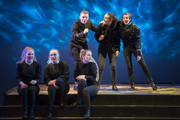 Various cast members of Scottish Operas Connect Chorus in Dido and Aeneas Scottish Opera 2017. They are standing and  sitting on stage singing with blue lights behind them.